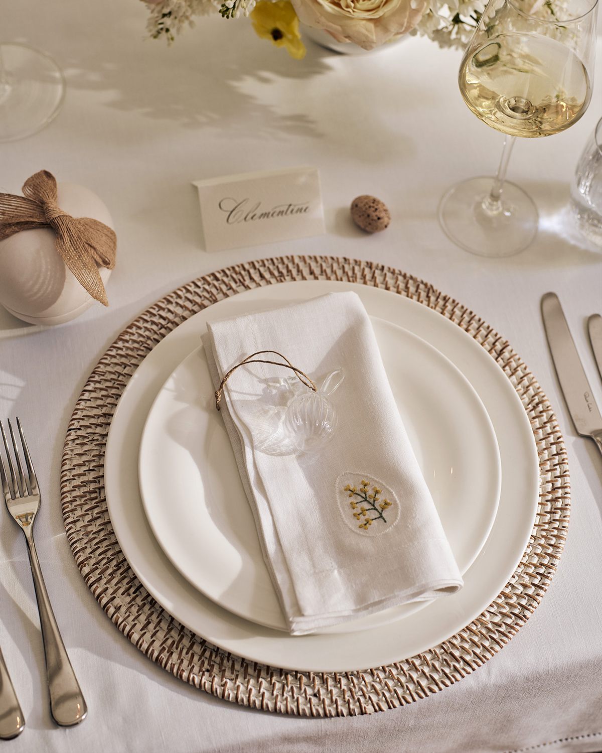 a simple easter tablescape with white tablecloth and plates and a name setting, with a white napkin with an embroidered easter egg and a glass bunny decoration on top of the plate