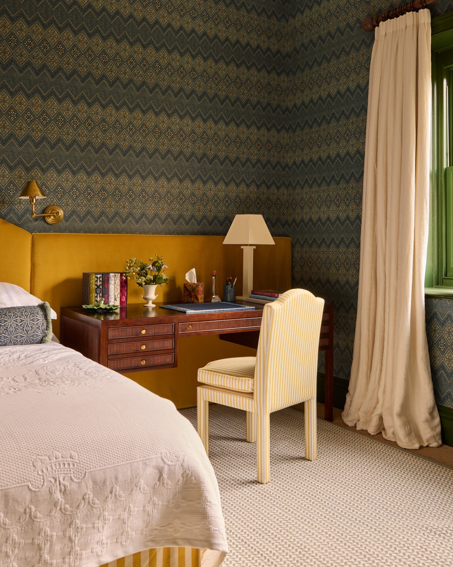 a bedroom with a blue and green zig zag patterned wallpaper, a large dark wooden desk with a yellow and white stripe chair and a bed with white bedding