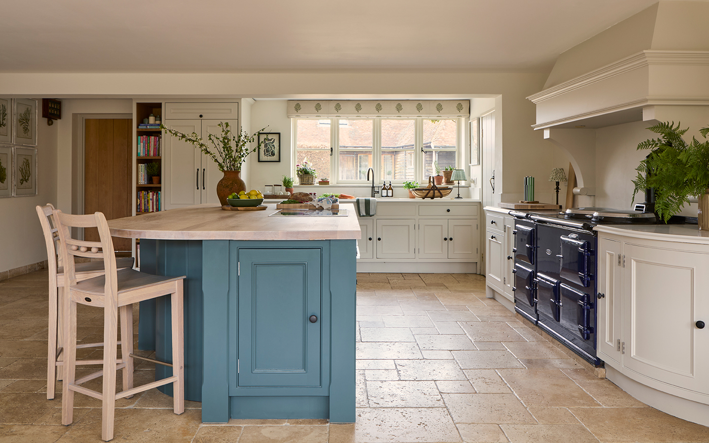 A kitchen with neutral cabinets topped with marble and a blue kitchen island topped with a wooden worktop. Wooden bar stools sit at the island. On the far wall is a large window dressed with a blind made in an artichoke-print fabric