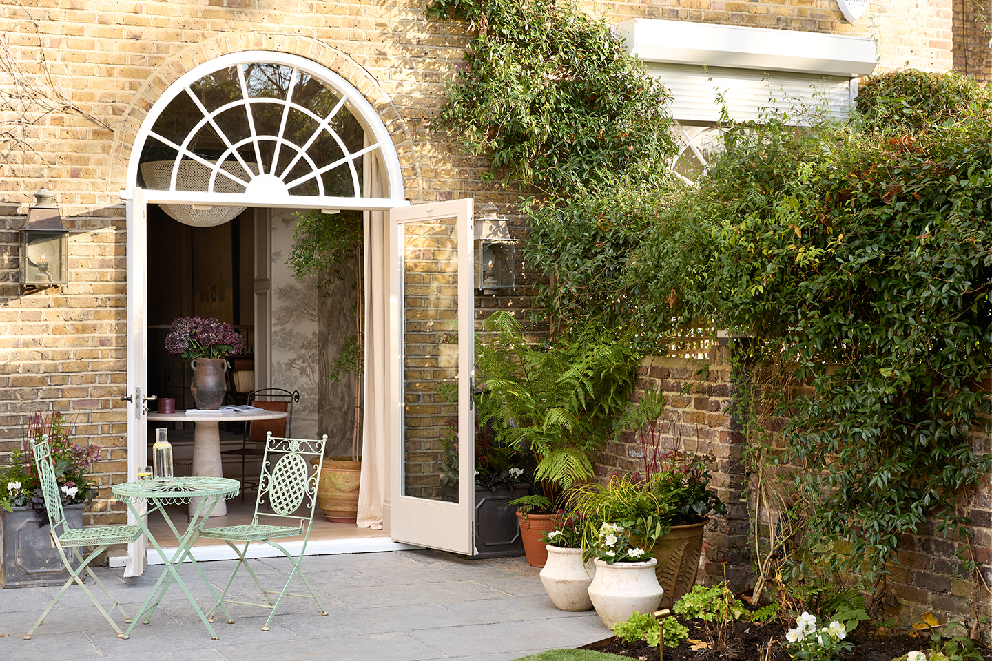 A townhouse garden including a flowerbed in the foreground, a brick wall draped in greenery to the right and a patio area with a green metal table and chairs. An arched double door leads through to a dining area inside
