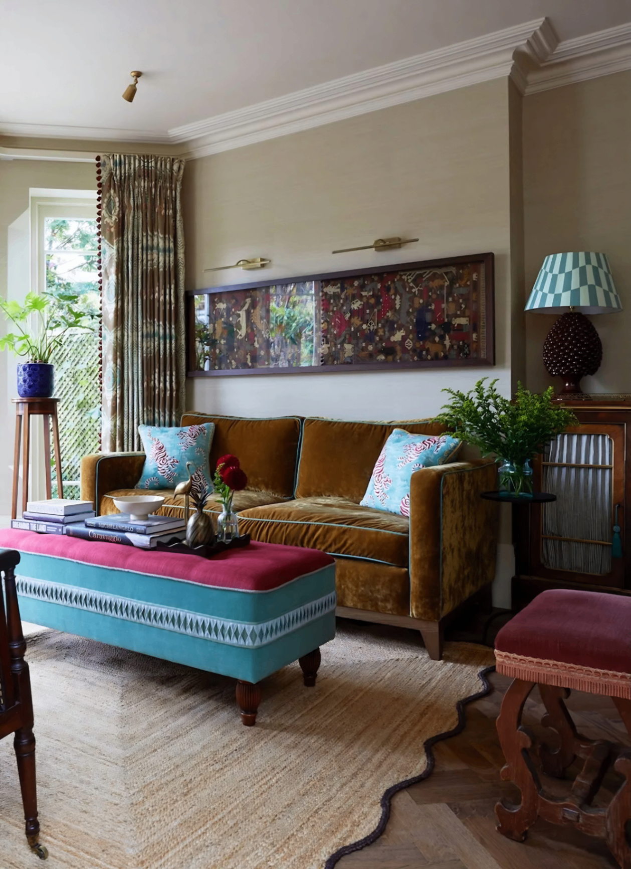A richly decorated living room featuring an ochre velvet sofa, a large teal and red fabric ottoman, and a textured natural rug on herringbone wood floors. Vintage furniture, pattern-rich textiles, and a long framed art piece add depth