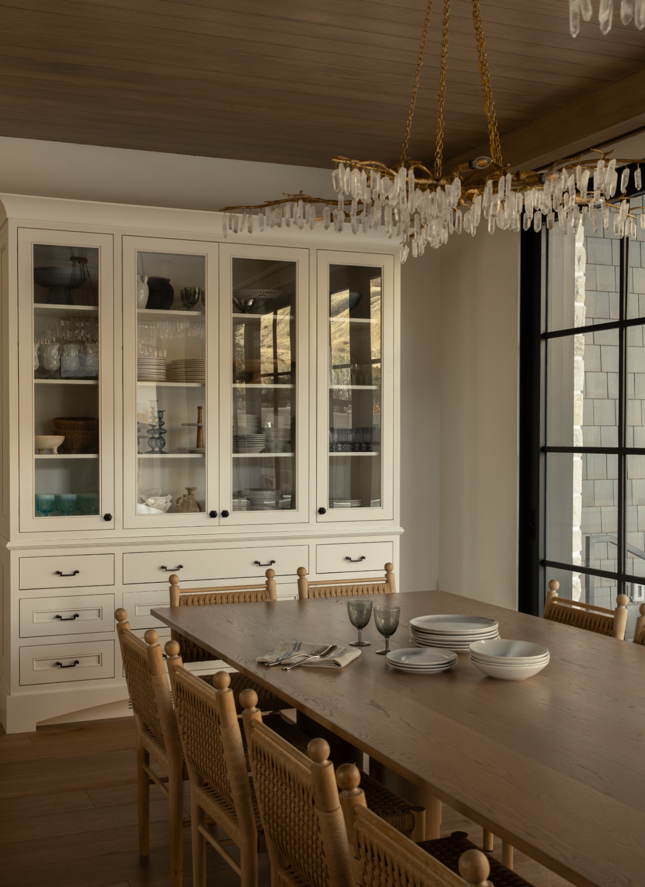 Elegant dining room with a long wooden table, woven seat chairs, a large cream fitted display cabinet, and a branch crystal chandelier