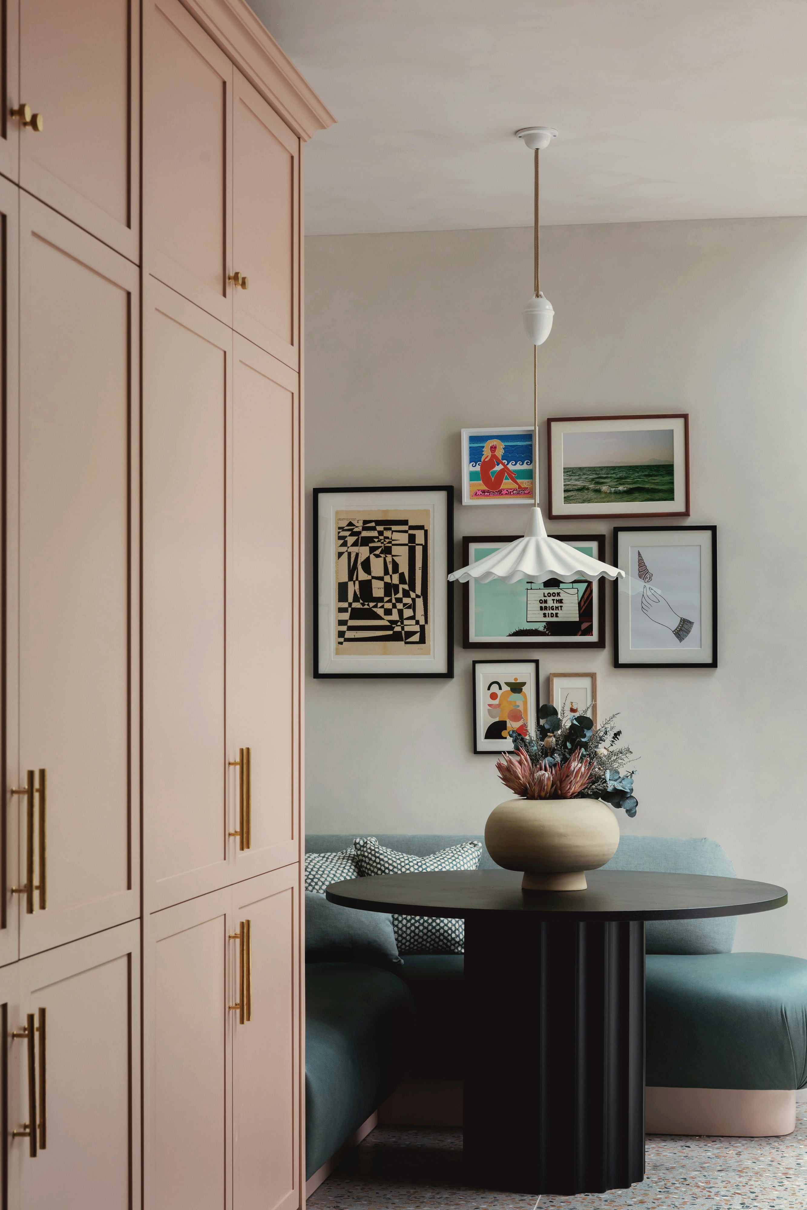 Modern small dining corner with a teal curved sofa booth, black ribbed pedestal table, pink cabinetry, and fluted pendant light