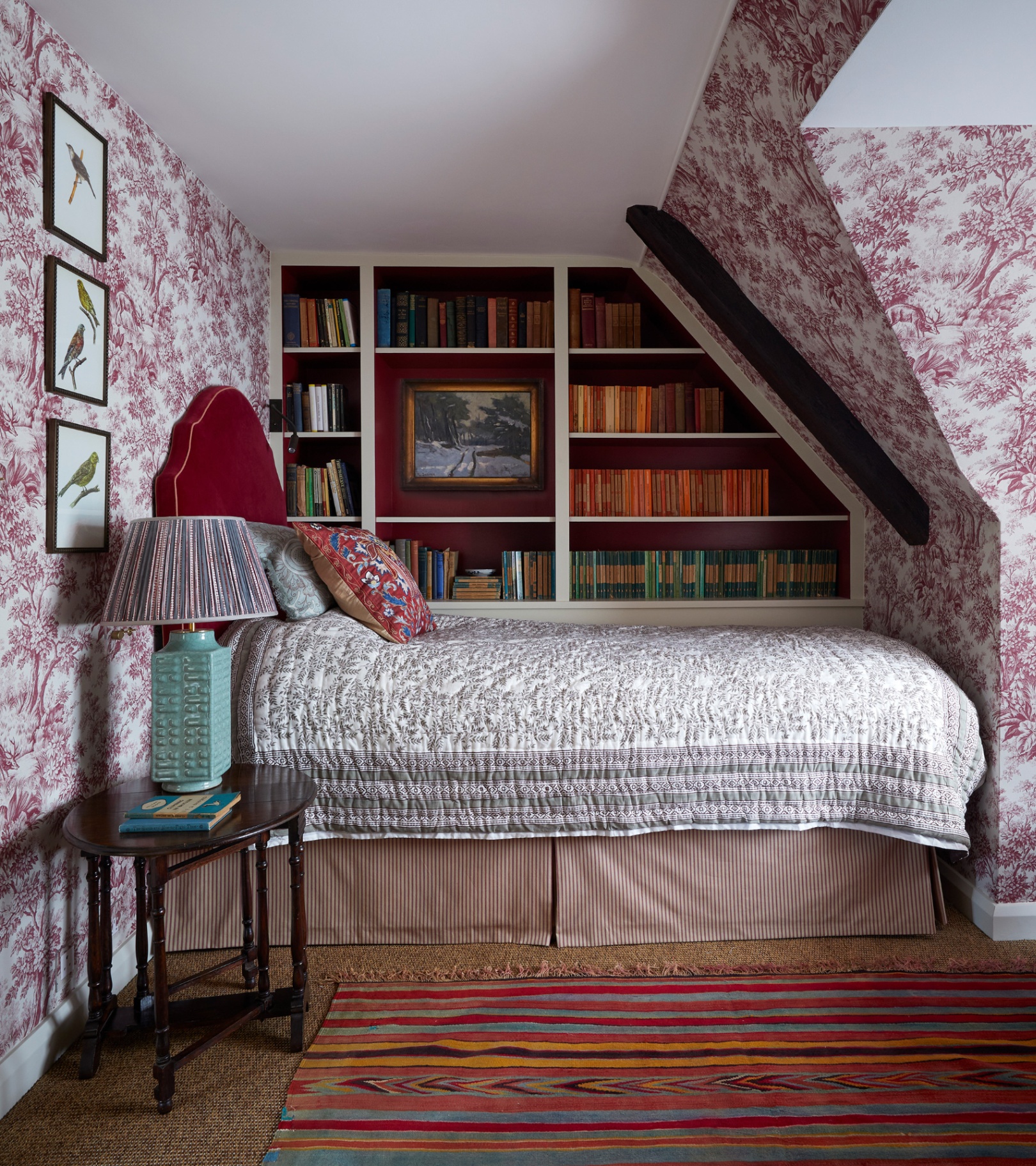 a bedroom with wooden beams and sloping ceiling with pink and white toile de jouy wallpaper a bed with white floral bedding and a built in bookcase on the back wall that is full of books