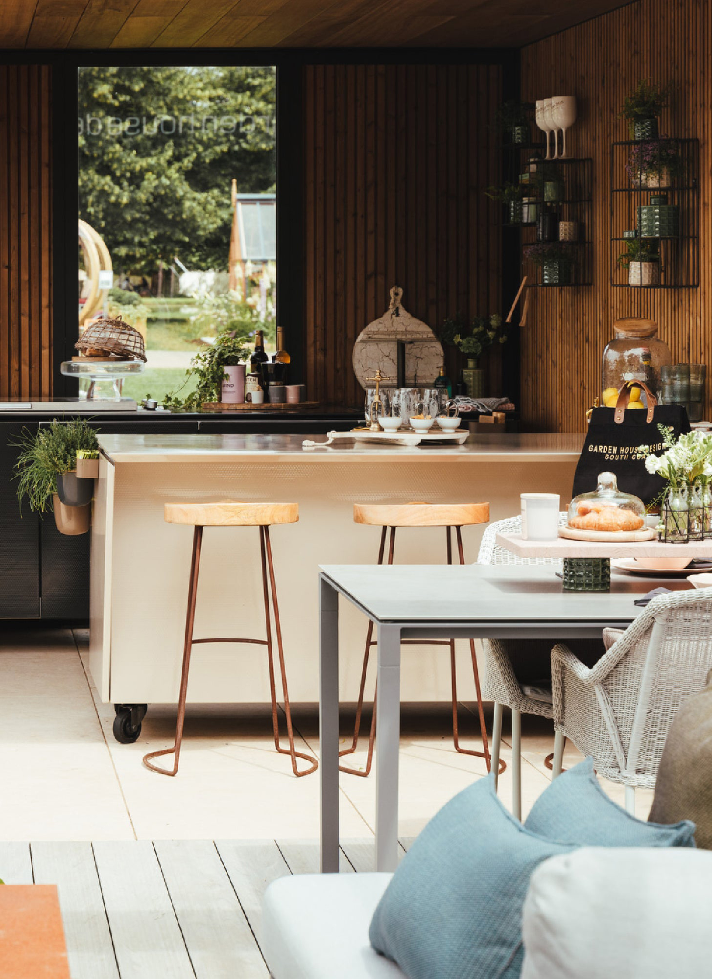 an outdoor kitchen area with wood panelled walls, a white counter with wooden stools and a grey dining table