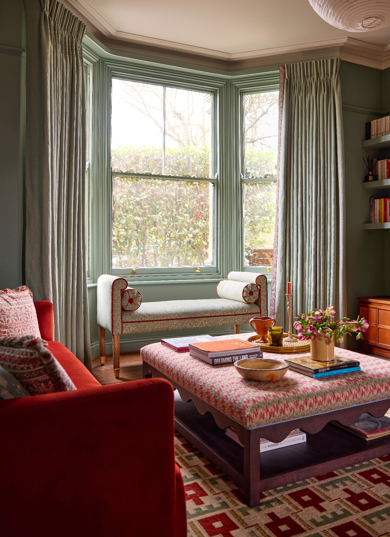 An elegant bay window sitting room featuring sage green walls framed by full-length sage drapes with red trim, an upholstered daybed bench, a vibrant red sofa, and a patterned ottoman coffee table.