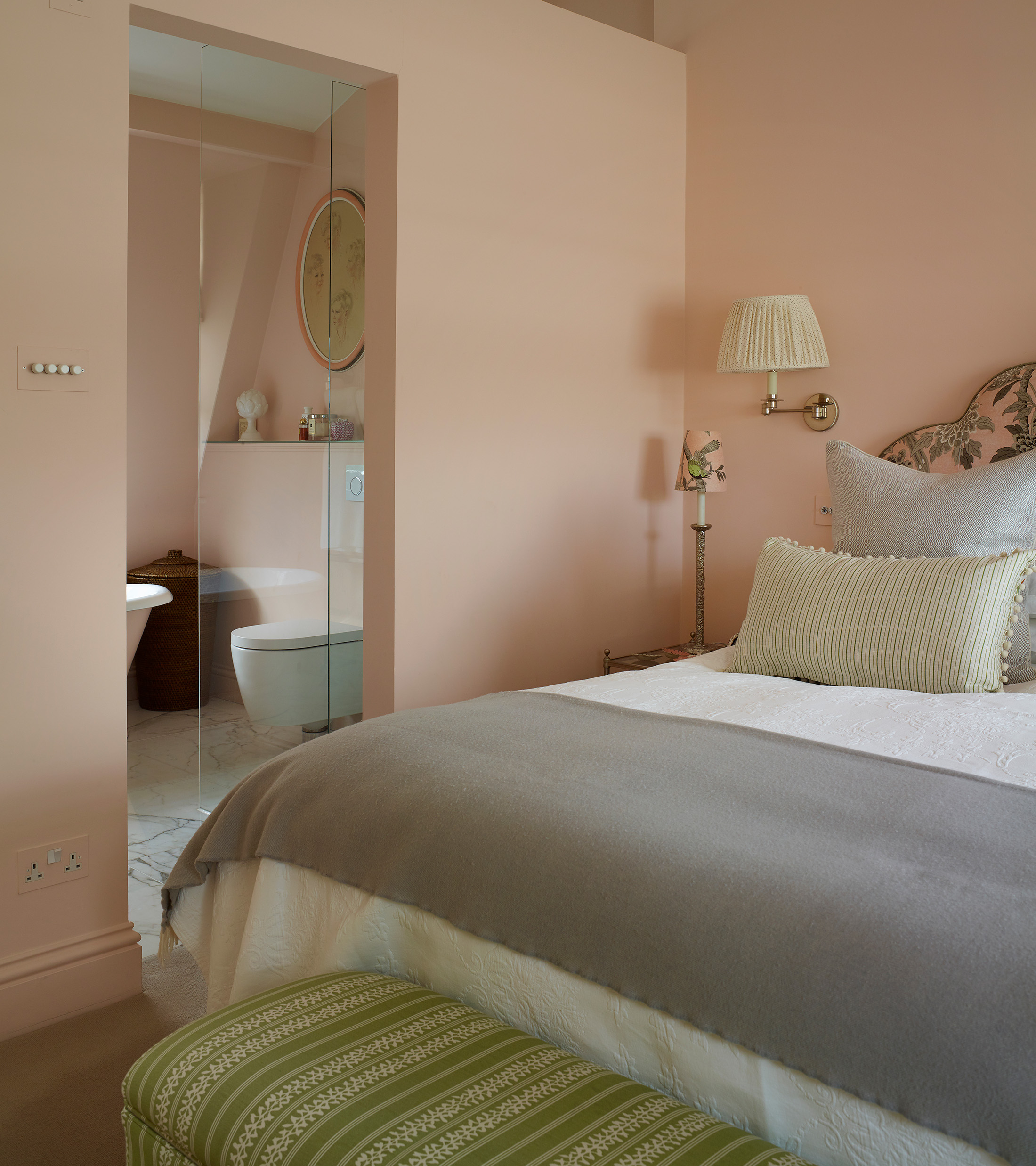 a pale pink bedroom looking through to the en suite, a bed with white bedding and a grey throw and a green ottoman at the foot of the bed