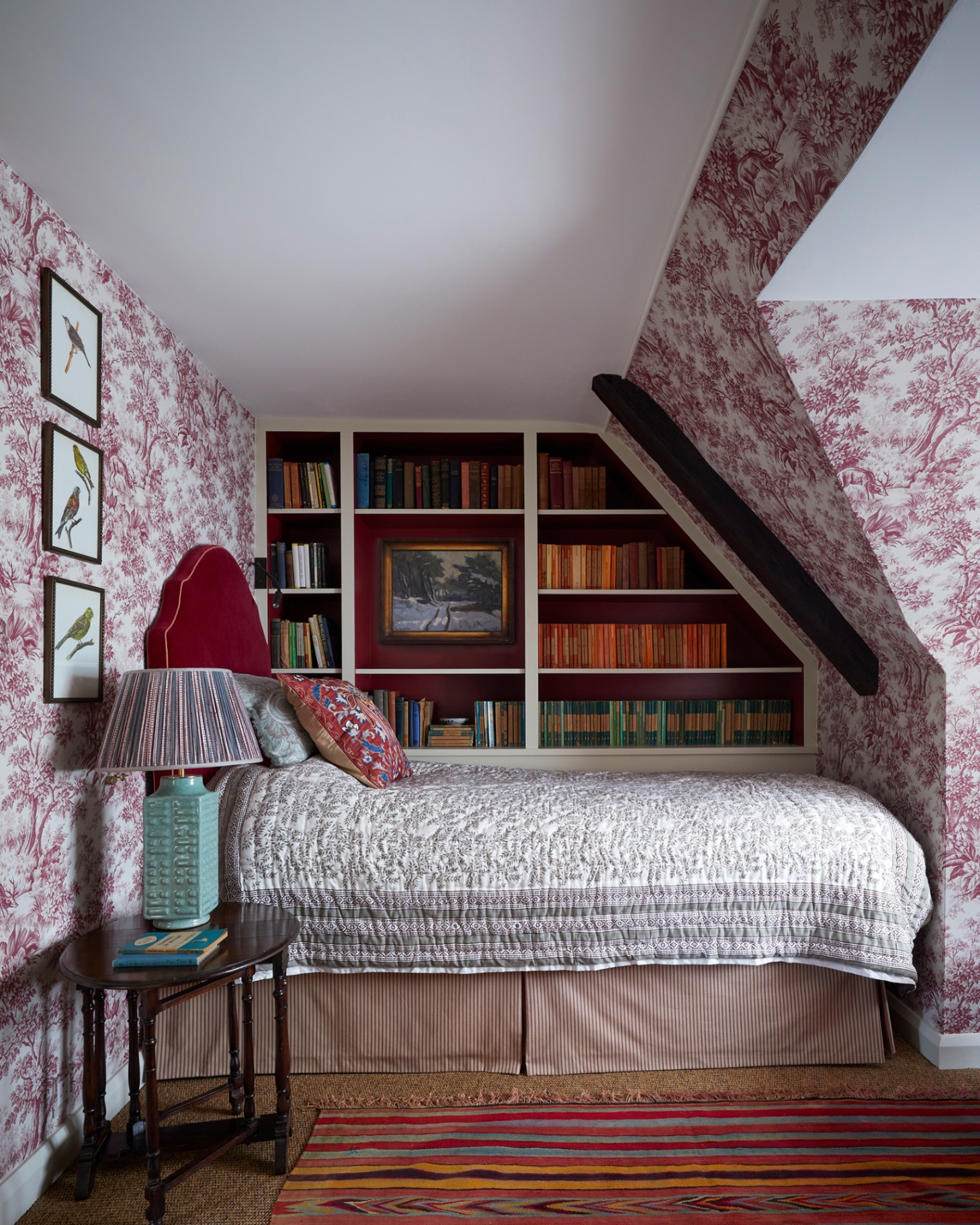 a cottage bedroom with sloping ceilings covered in a red and white patterned wallpaper, a library wall behind a single bed and a bedside table with a large bedside table lamp