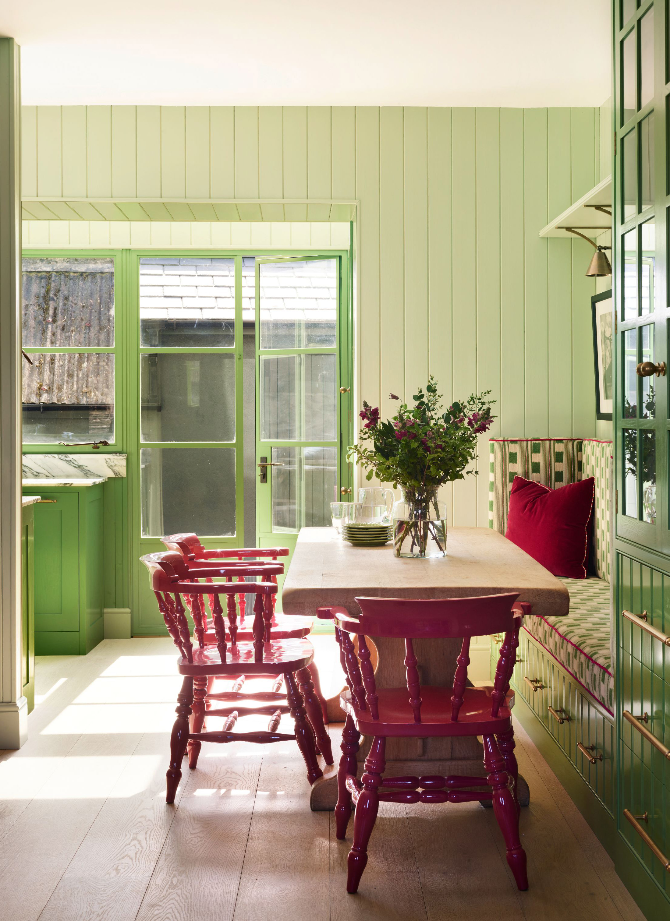 Lime green wood paneled dining nook with a light wood table, high-gloss magenta spindle chairs, and a patterned banquette