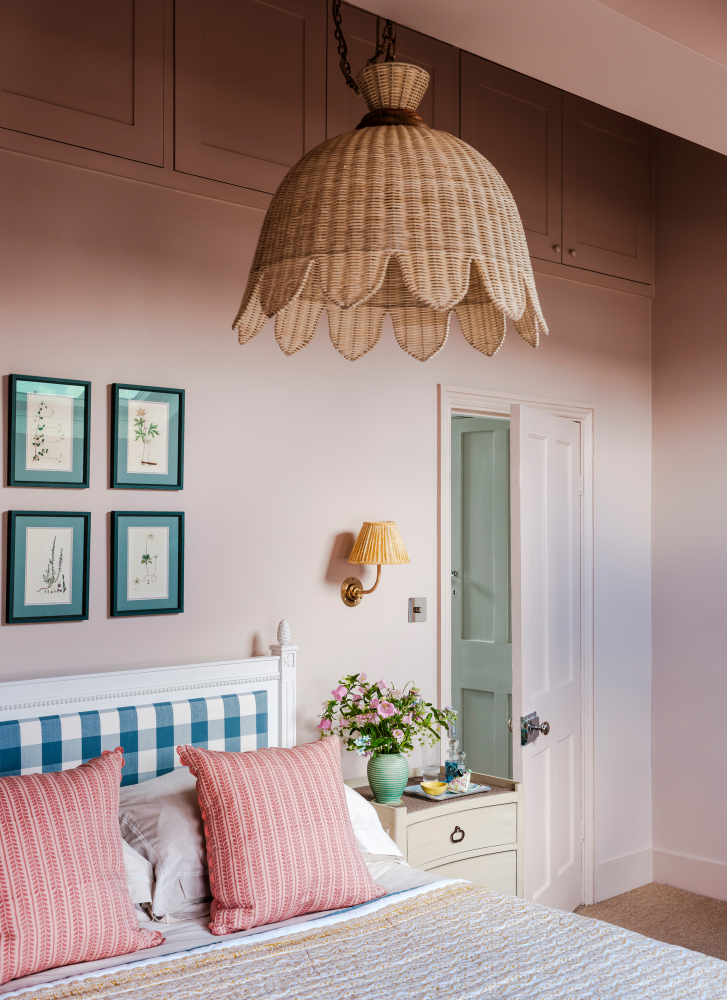a bedroom with pale pink walls and door, a large rattan pendant shade, a yellow sconce and a bed with a gingham headboard and pink pillows