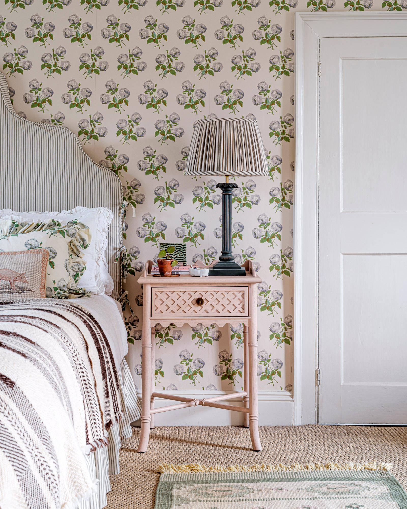 a bedroom with white and blue floral wallpaper, a pale pink bedside table with a large black and white stripe pleated table lamp and a bed with a stripe headboard and a stripe bedspread