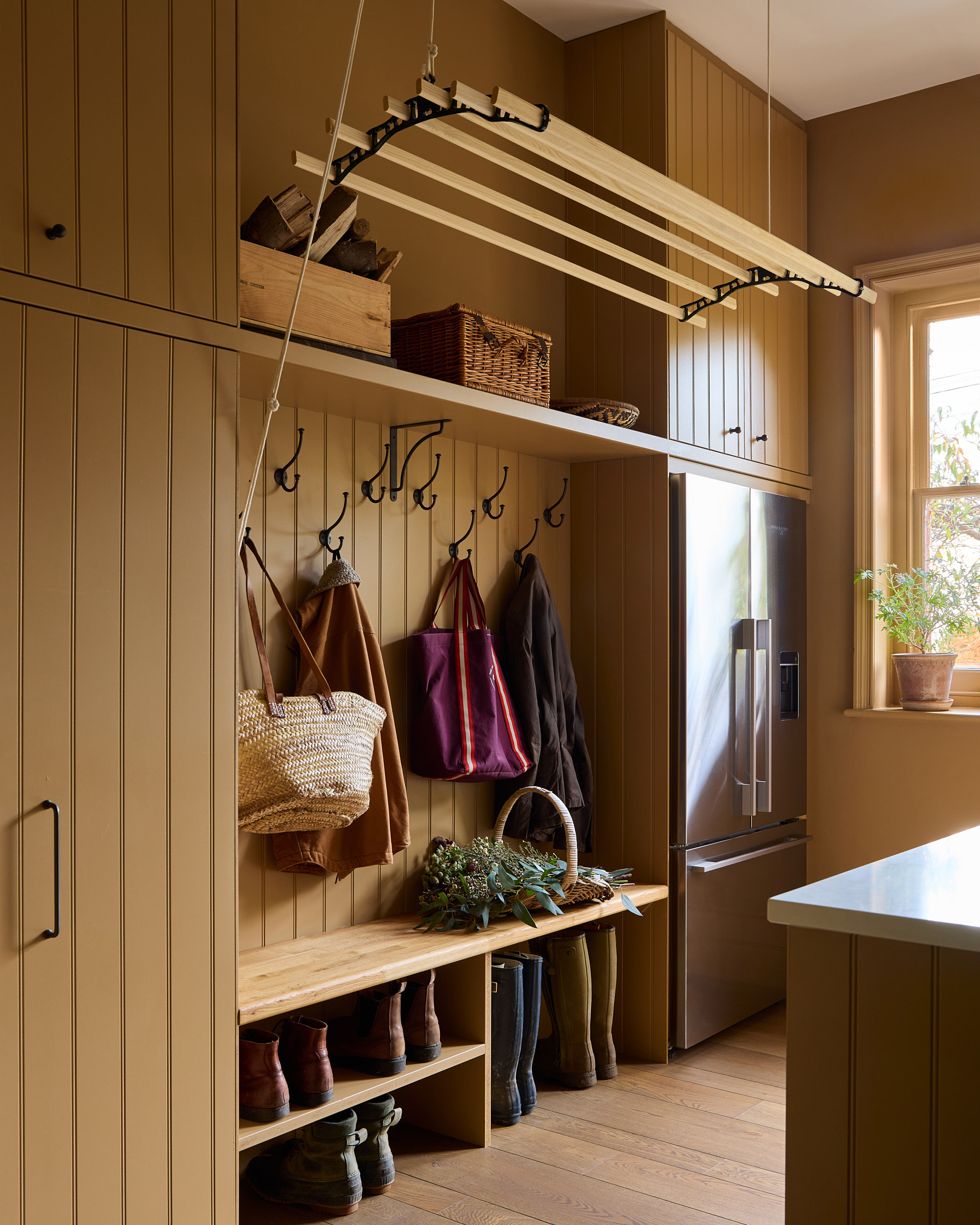 a mustard yellow painted utility and boot room with a wooden hanging clothes rack overhead, a clothes peg and boot station and a large silver fridge