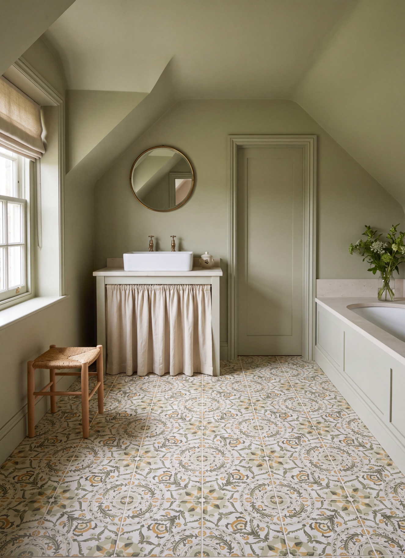 Sage green bathroom featuring a skirted vanity with a vessel sink and circular mirror beneath sloped ceilings. Patterned floor tiles span the room toward a built-in bathtub, wooden stool, and natural window light