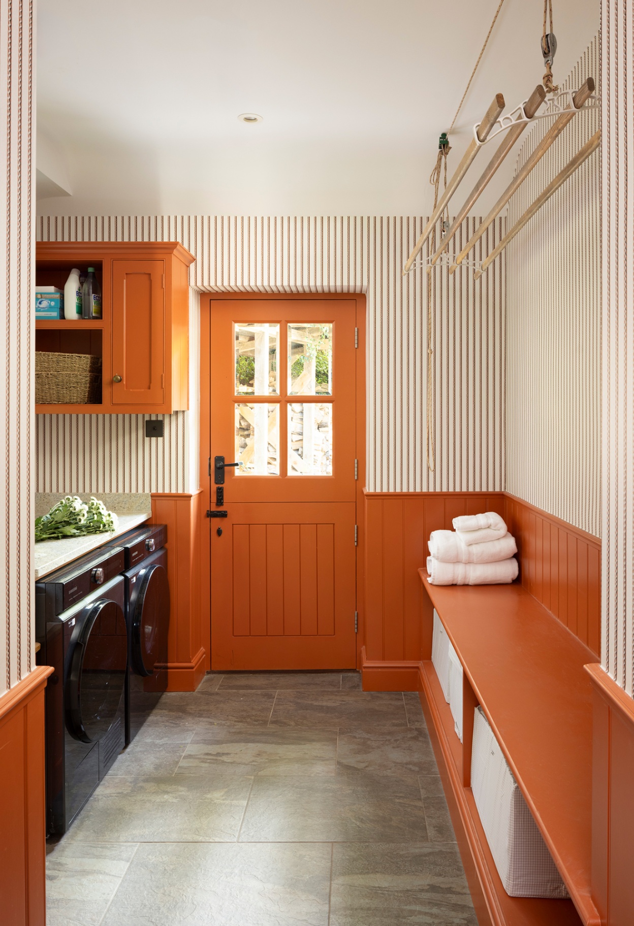 a utility room with a black washing machine and dryer on the left and a built in bench on the right. The walls are papered in a simple stripe on the upper half and painted orange tongue and groove on the lower half, with an orange painted cabinet and door