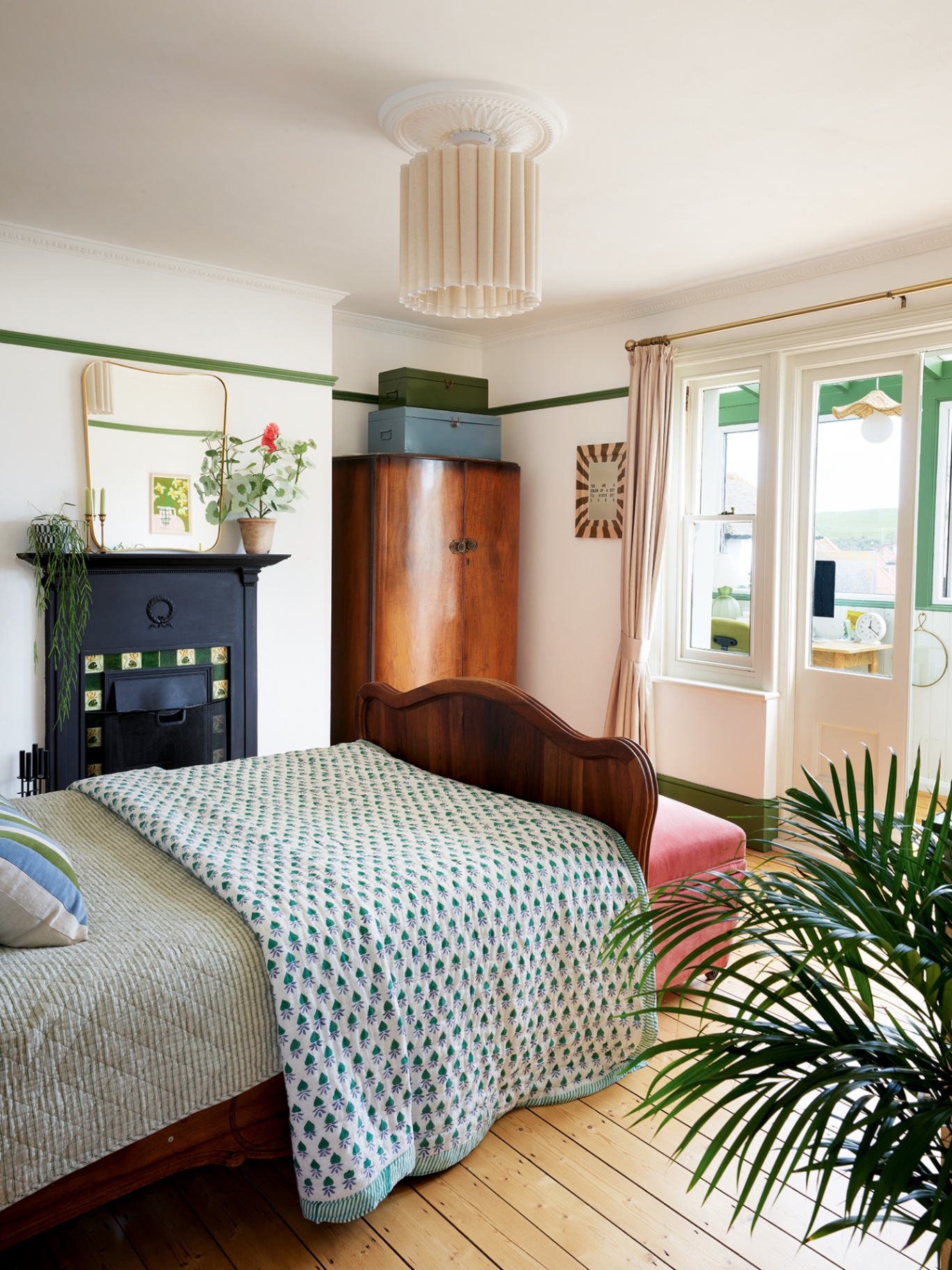 An angled shot of a bedroom with a wooden double bed dressed with striped linens and a block-print throw in the foreground. In the background is a large wooden wardbrobe, black fireplace and a door leading onto a small enclosed balcony