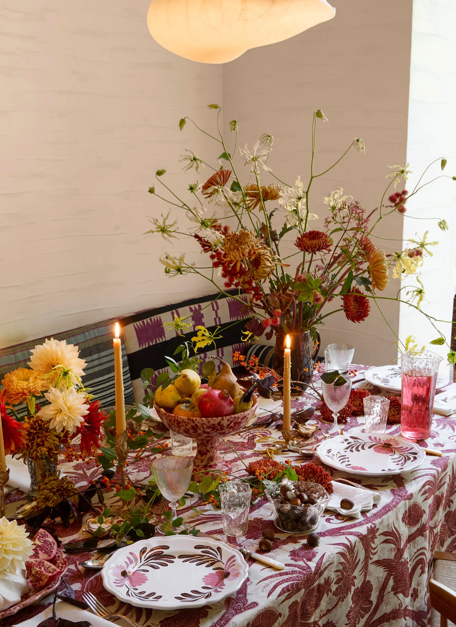 an autumn table with a white nad dark pink floral tablecloth, vases of white, red and orange dahlias, a footed fruit bowl, white and pink floral plates and yellow dinner candles