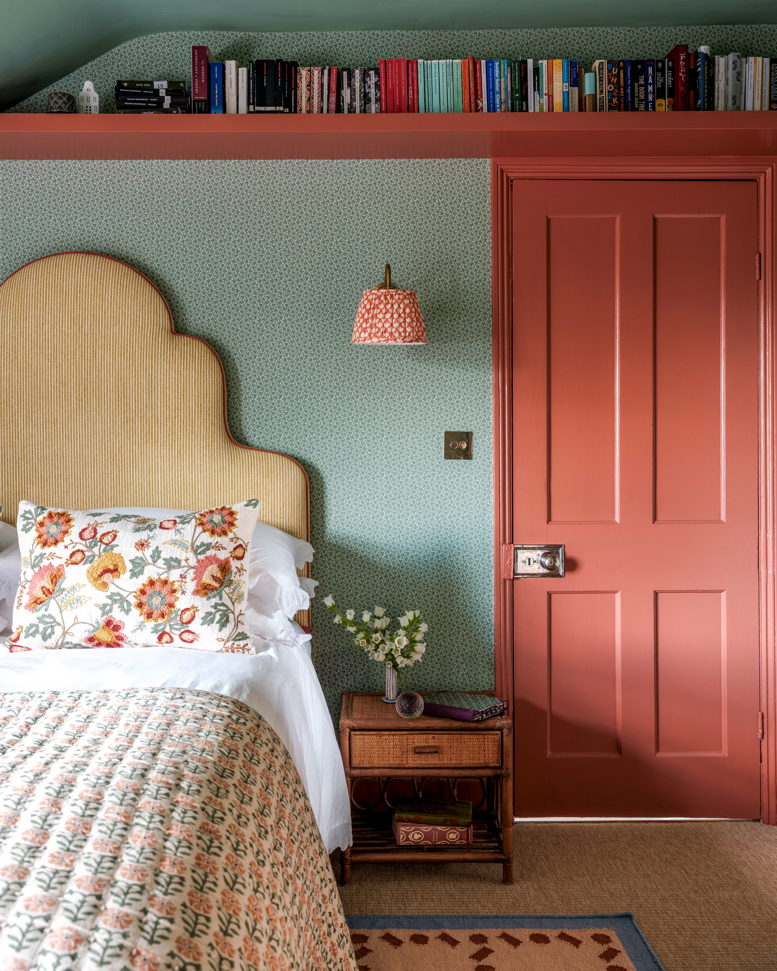 a bedroom with coral pink door and matching high perimiter shelf filled with colourful books, a green patterned wallpaper and a bed with a large yellow striped upholstered headboard with a floral pillow and bedspread