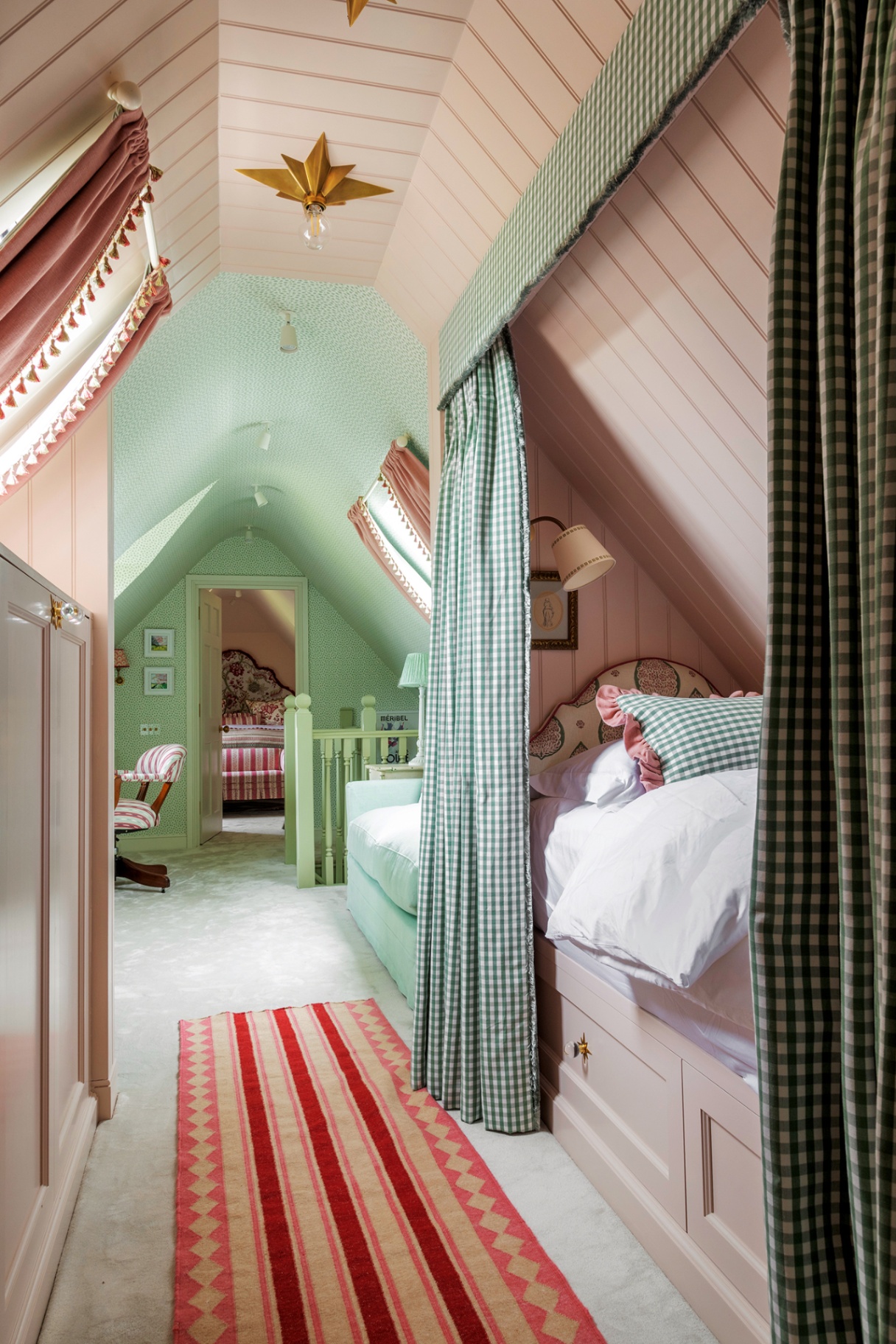 an attic bedroom with pink tongue-and-groove walls and ceiling in the foreground and a built-in single bed nook, and pale green walls and ceiling and a sofa and desk with chair in the background. A door leads through to a double bedroom at the back