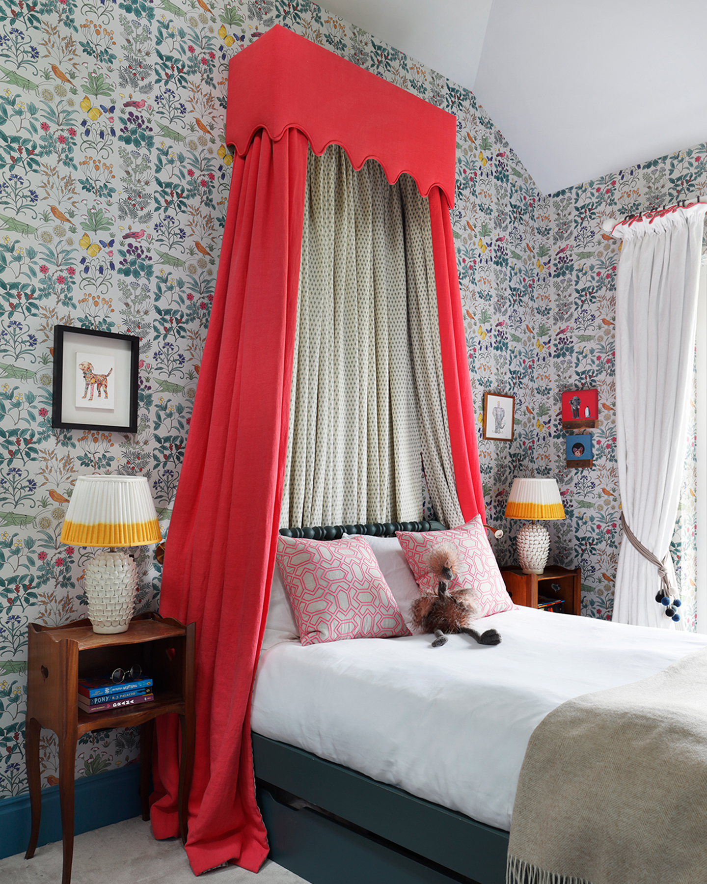 a children's bedroom with colourful botanical wallpaper, a bright red bed canopy over a single bed with white bedding a a stuffed toy and two dark wood bedside tables with white and yellow lamps on top
