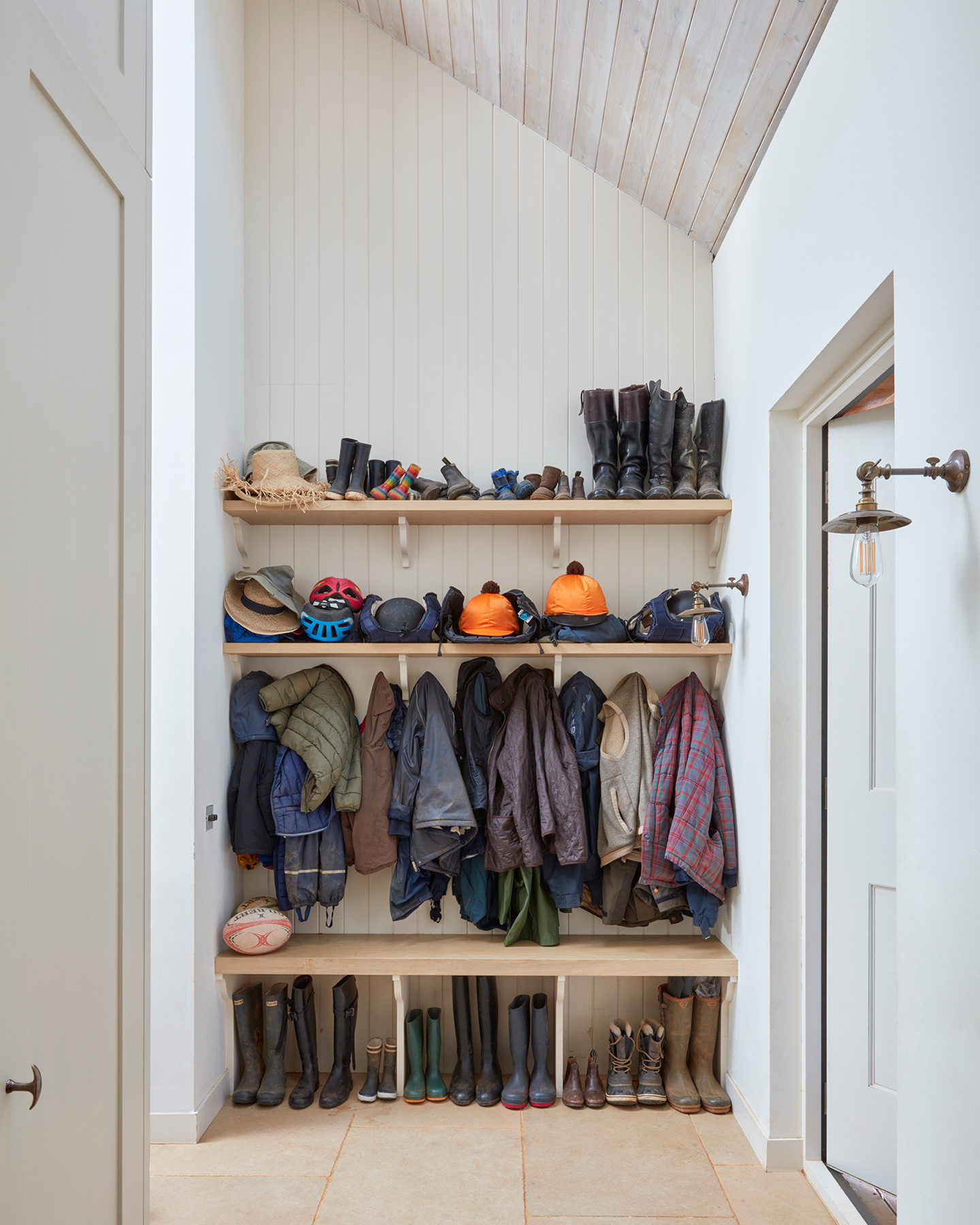 A high-ceilinged boot room area with timber-clad walls and angled ceiling. Wooden shelves and racks are built in and store an array of boots, hats and coats