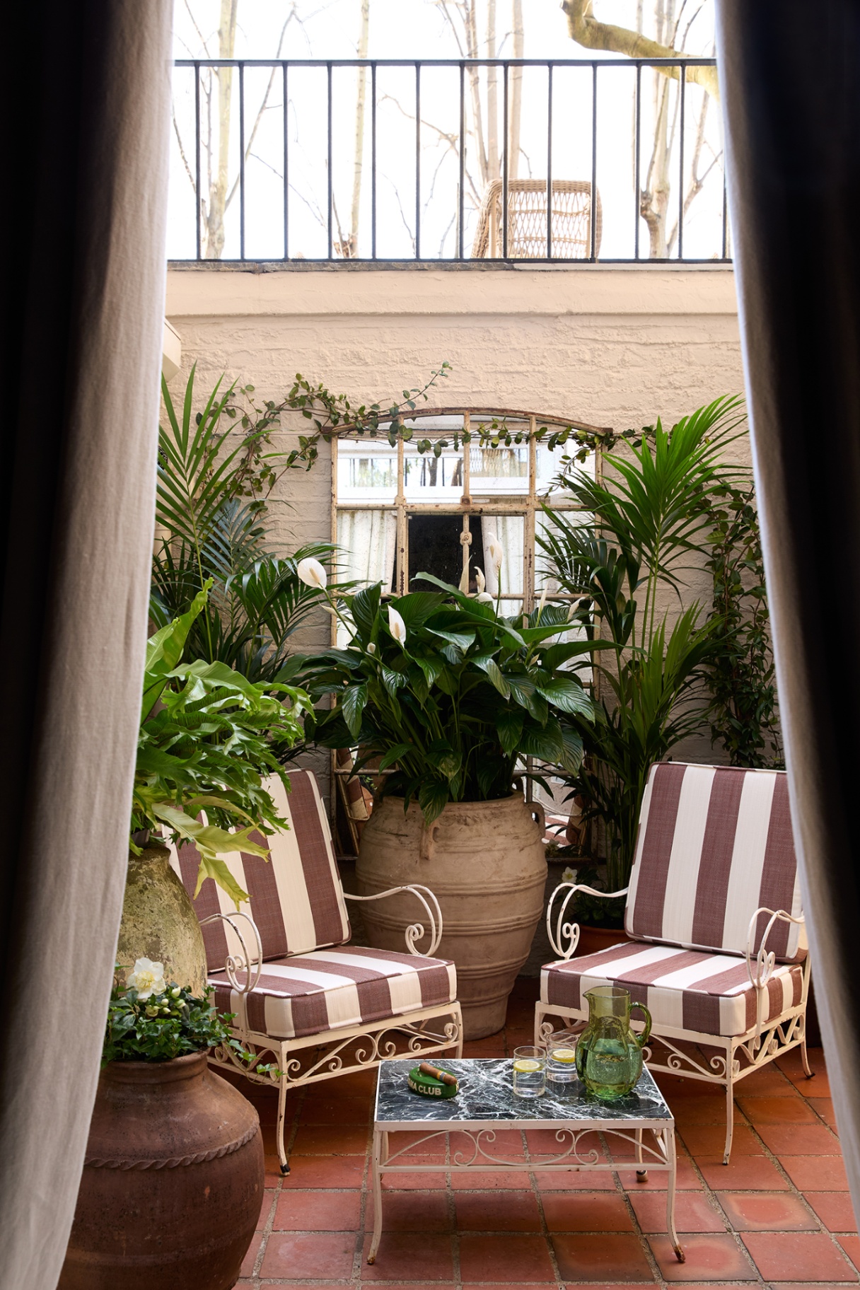 two garden chairs with brown-and-white striped cushions and a small coffee table topped with a jug and glassware are arranged on a courtyard filled with pots and greenery