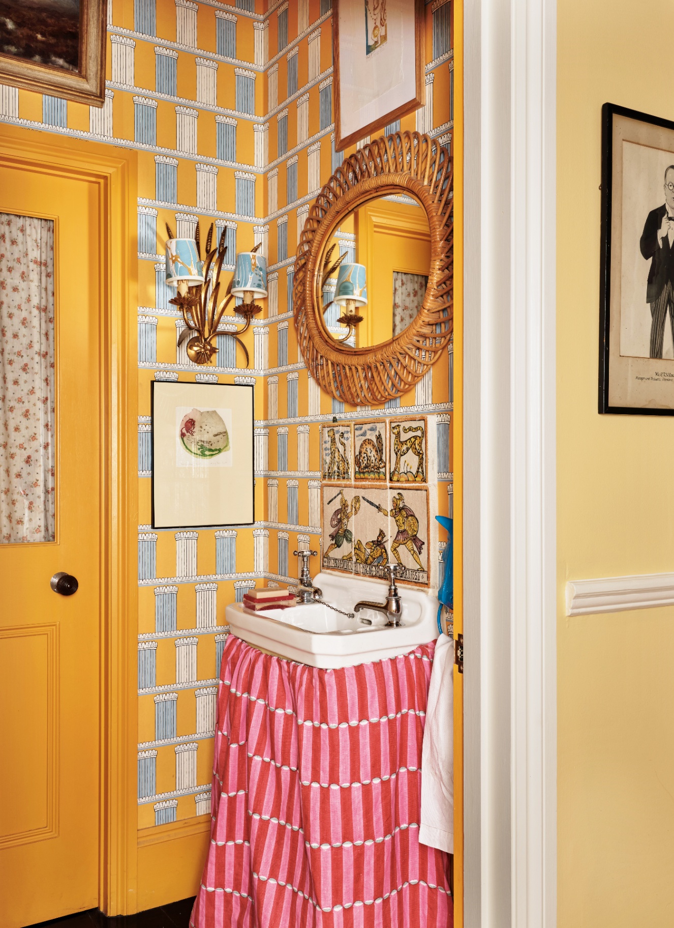 a downstairs toilet with bright yellow wallpaper with pale blue and white patterns, a bright yellow door, a pinka nd red patterned sink skirt, a patterned tile splashback, a rattan mirror and a double sconce with pale blue shades