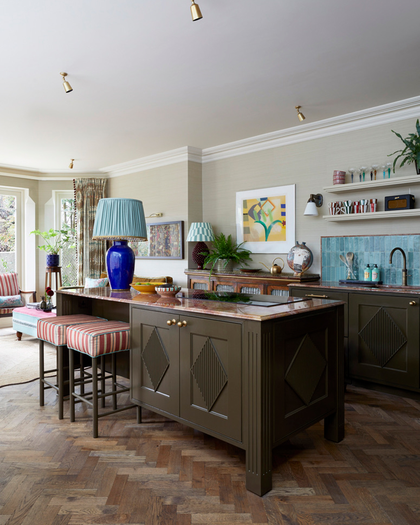 a kitchen with deep brown painted cabinets and kitchen island, a bright blue tiled splashback, parquet flooring and a large electric blue lamp with alight blue pleated lampshade