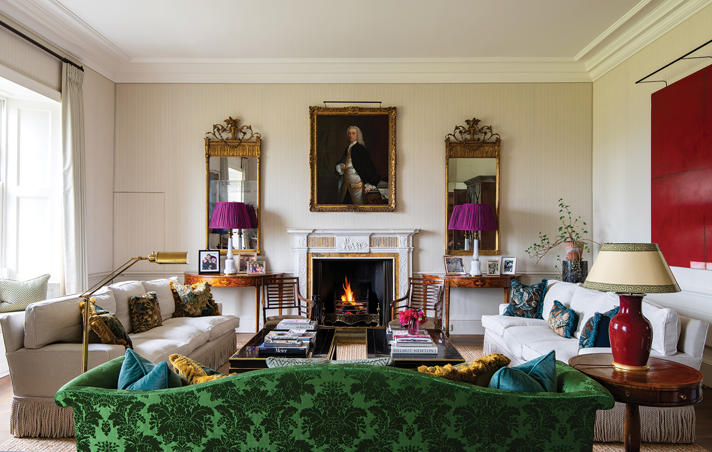 a drawing room with a patterned velvet green sofa in the foreground and two fringed white sofas, one left and one right of a pair of black lacquer coffee tables. In the centre of the back wall is a fireplace with a lit fire and a portrait hanging above