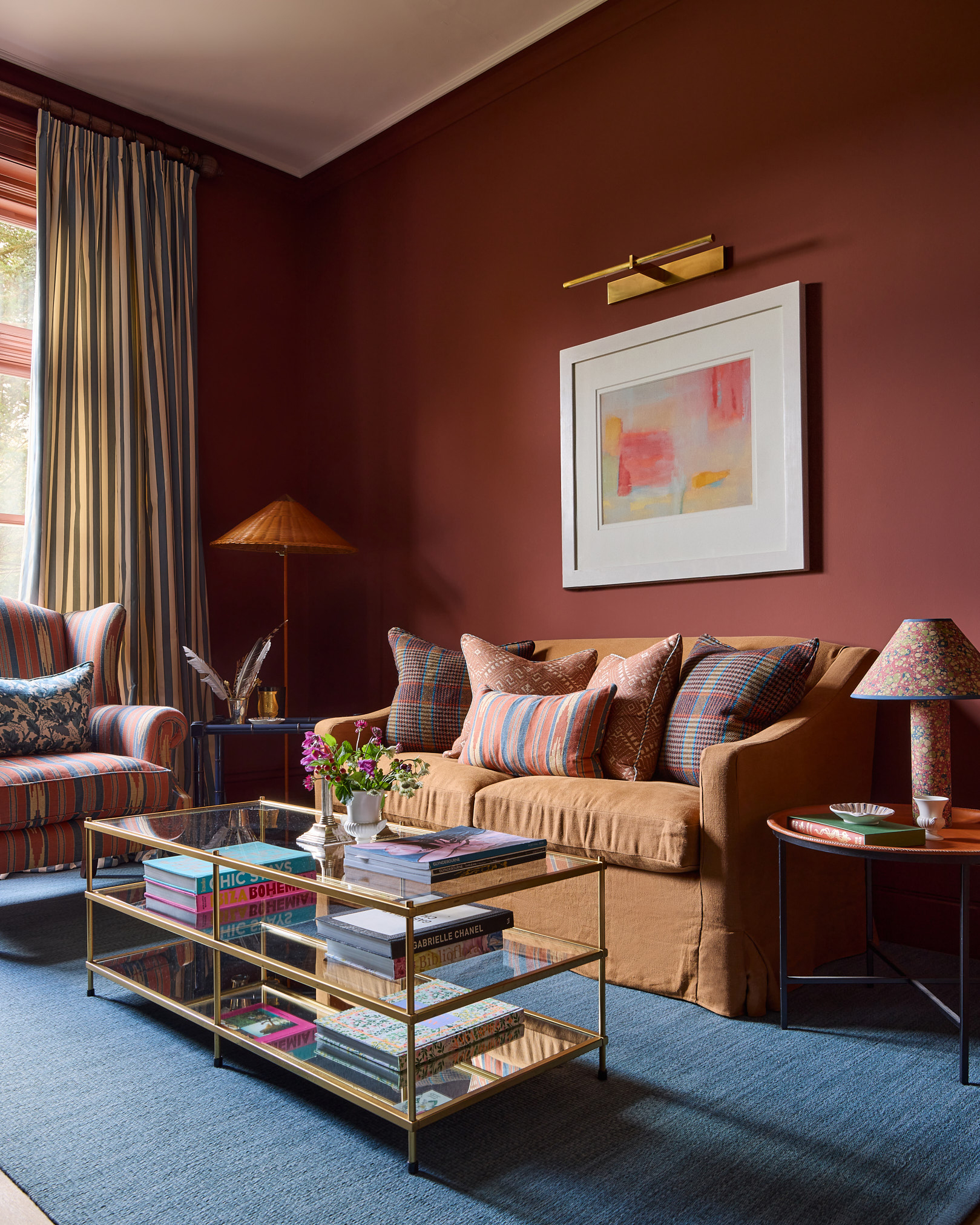 a living room with warm red walls, a mustard yellow sofa with patterned cushions, a gold glass coffee table full of books and white and blue stripe curtains