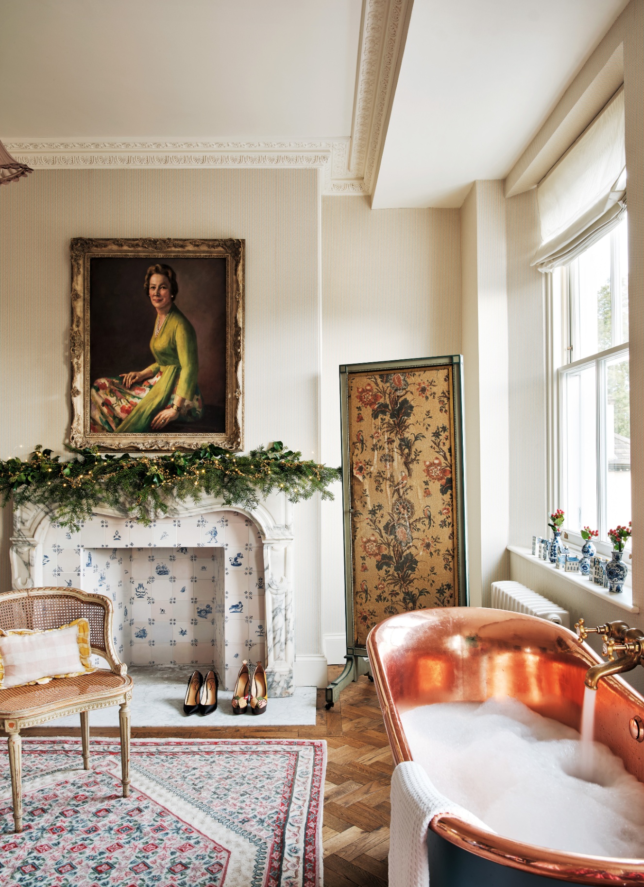 A luxurious bathroom featuring a copper roll-top tub filled with bubbles alongside a tiled decorative fireplace draped with a lit Christmas garland. A large oil portrait hangs above the mantel, set against patterned wallpaper and parquet flooring