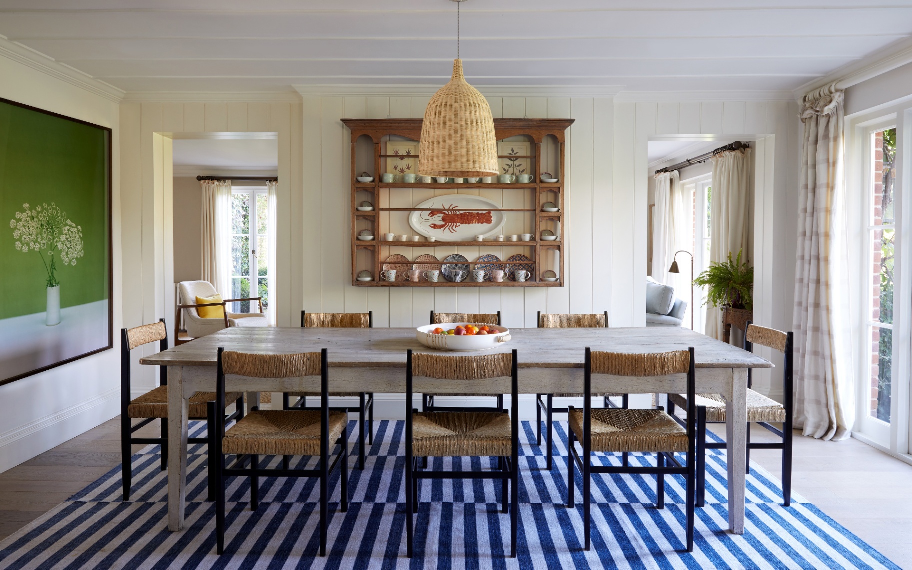 a dining room with a large white washed wooden dining table, rattan dining chairs, a blue and white srtiped rug, a large open shelving unit hung on the wall with a red lobster plate and a rattan pendant light