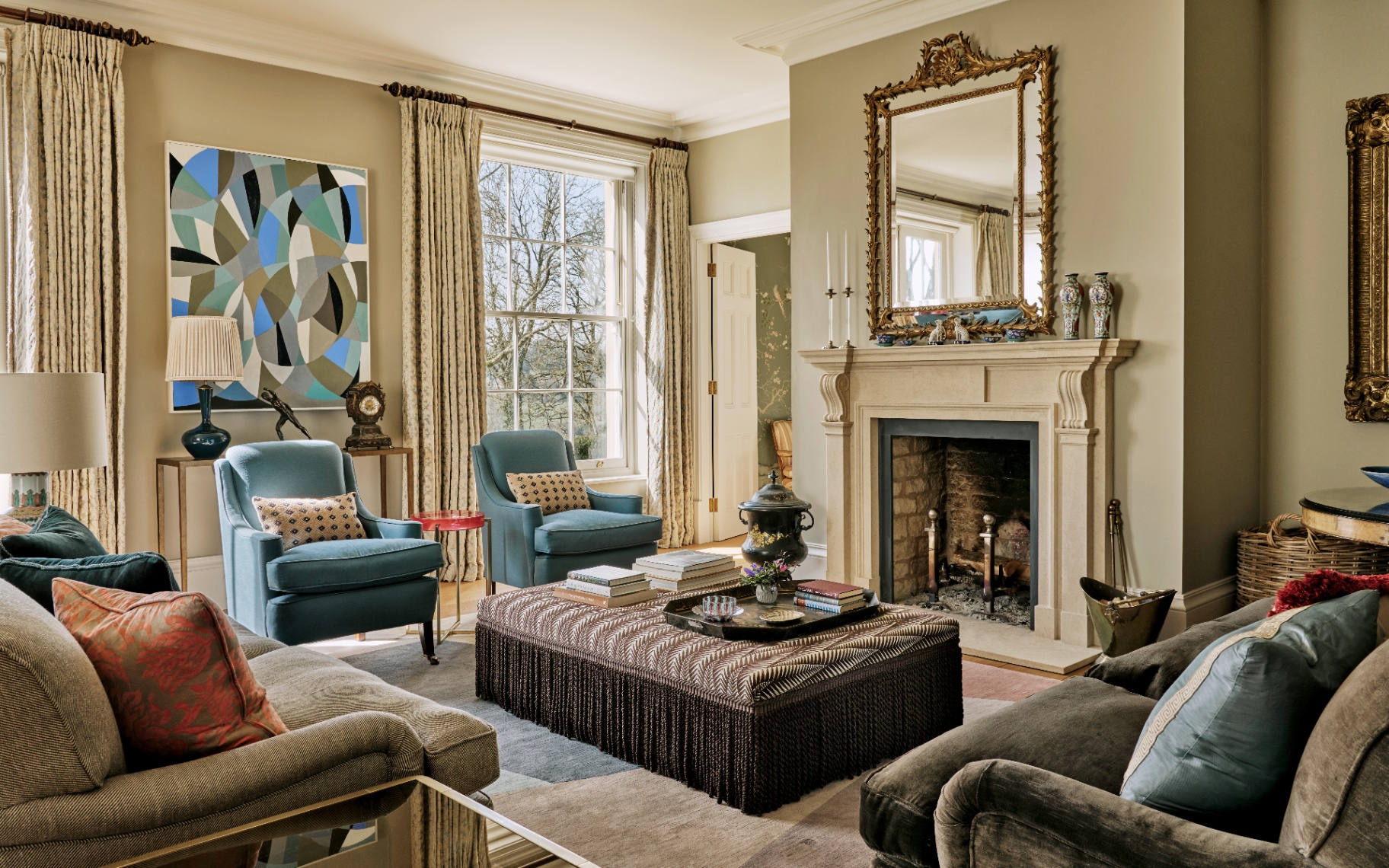 a living room with pale green walls, two blue armchairs, a brown patterned ottoman with fringing, a large fireplace with a large gol ornate mirror above