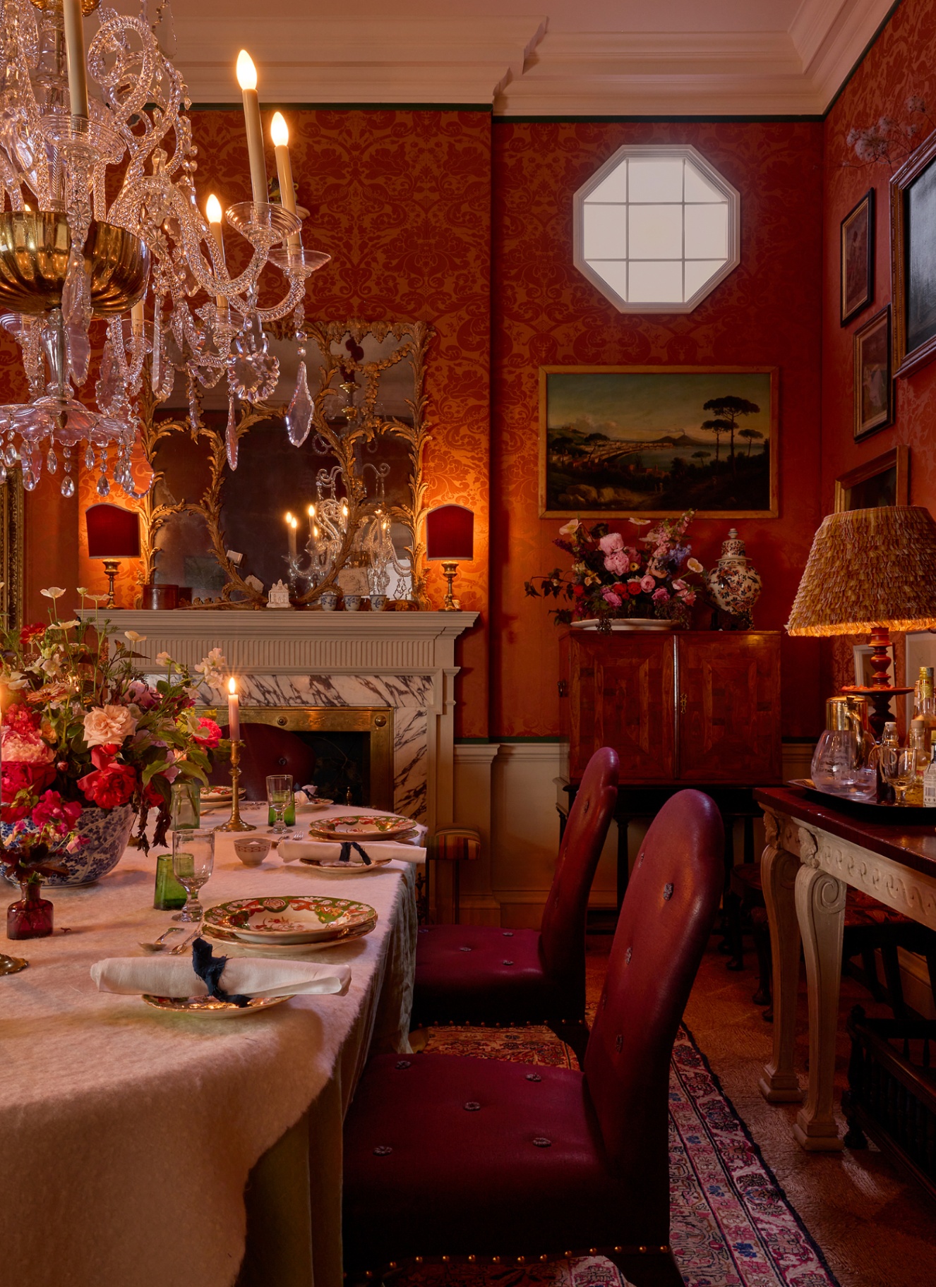 Atmospheric red dining room featuring an ornate crystal chandelier above a set dining table, rich Damask wallpaper, and warm glowing lamps