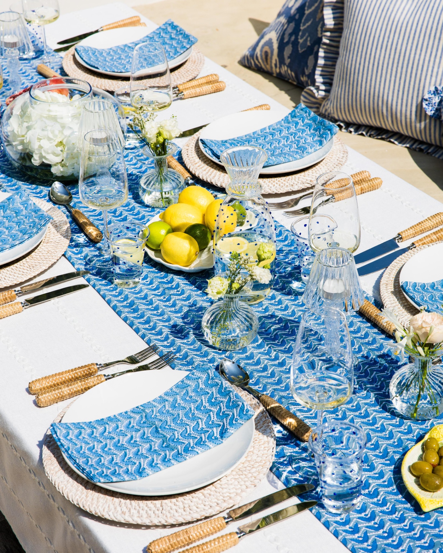 A summer tablescape with a blue patterned runner and matching napkins, cutlery with rattan hadnles and a bowl of lemons in the middle