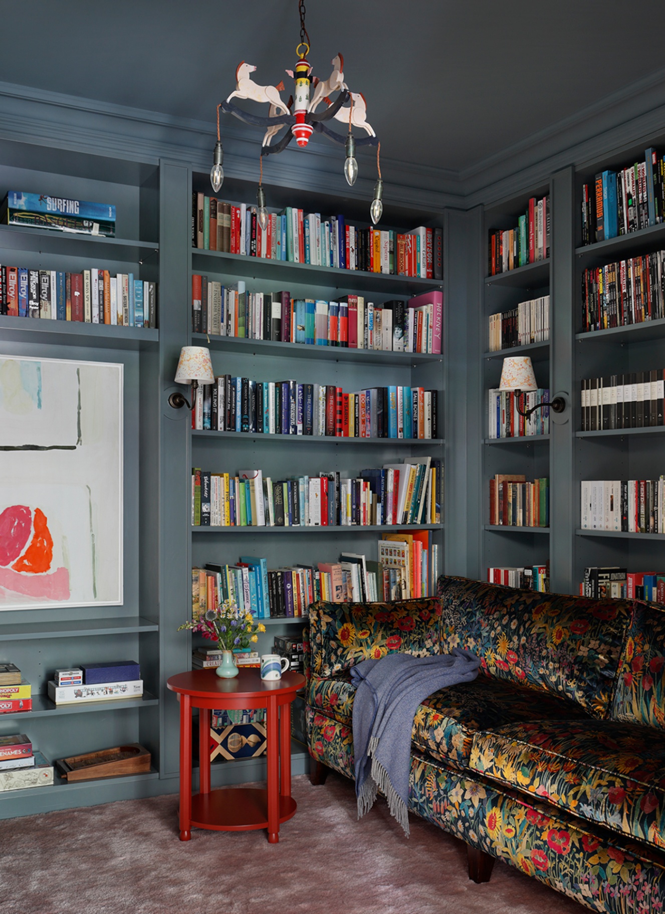 a living room corner wtih a dark floral velvet sofa, a bookcase wall painted in blue filled with colourful books and a small red side table