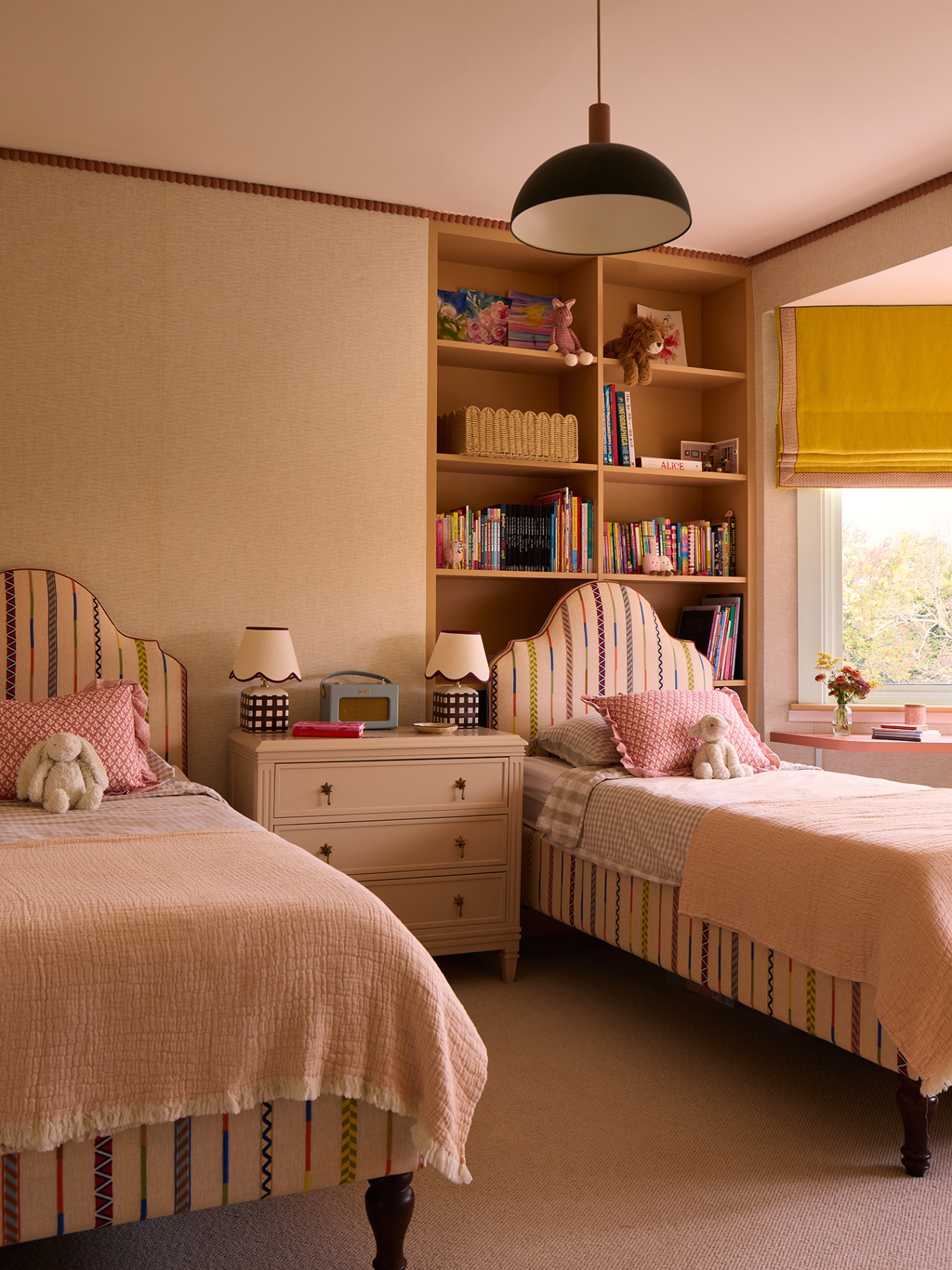 A children's bedroom with striped upholstered twin beds separated by a chest of drawers topped with table lamps and a radio. Built in bookshelves hold books and toys and a pendant light hangs from the ceiling. A yellow and pink blind dressed the window