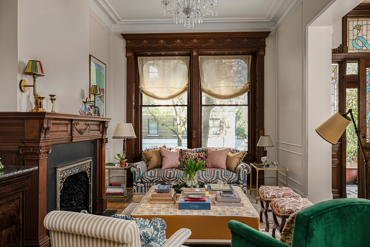 A living room with taupe walls and a large double window in the back dressed with relaxed yellow linen blinds. A patterned sofa, striped armchair, velvet armchair and two stools are arranged around an upholstered ottoman. A large fireplace sits to the left
