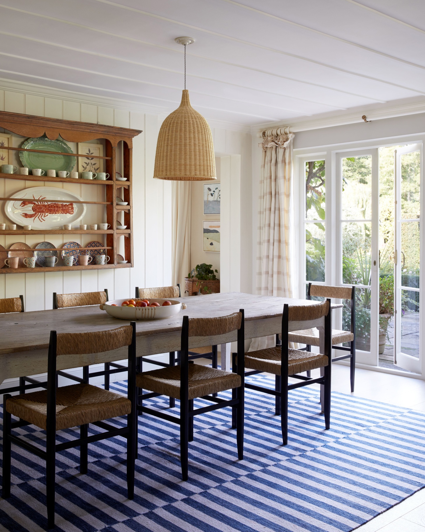 a dining room with a blue and white stripe rug underneath a large wooden dining table with rattan dining chairs, a rattan pendant light and a large open shelving unit on the wall with an oval lobster plate