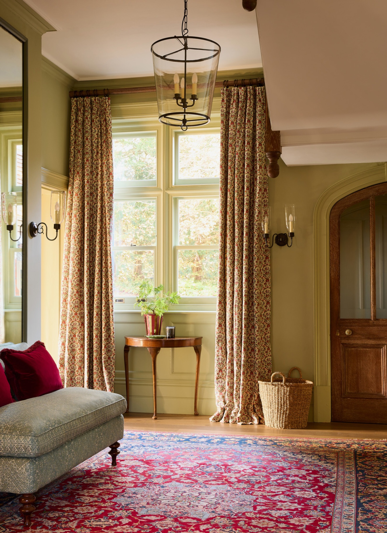 a hallway with high ceilings, sage green walls, a red patterned rug a large window with sage and red floral curtains and a large wooden door