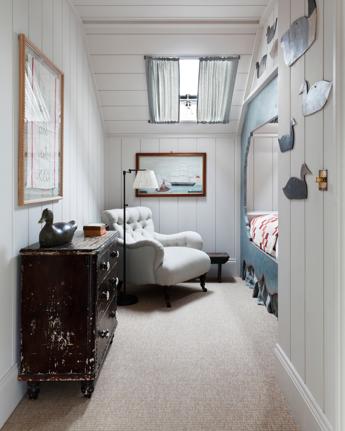 a bedroom with a skylight covered in a blue check curtain, a jute stripe carpet, an old dark wood chest of drawers, a white and blue stripe armchair and a painting of a sail boat above it