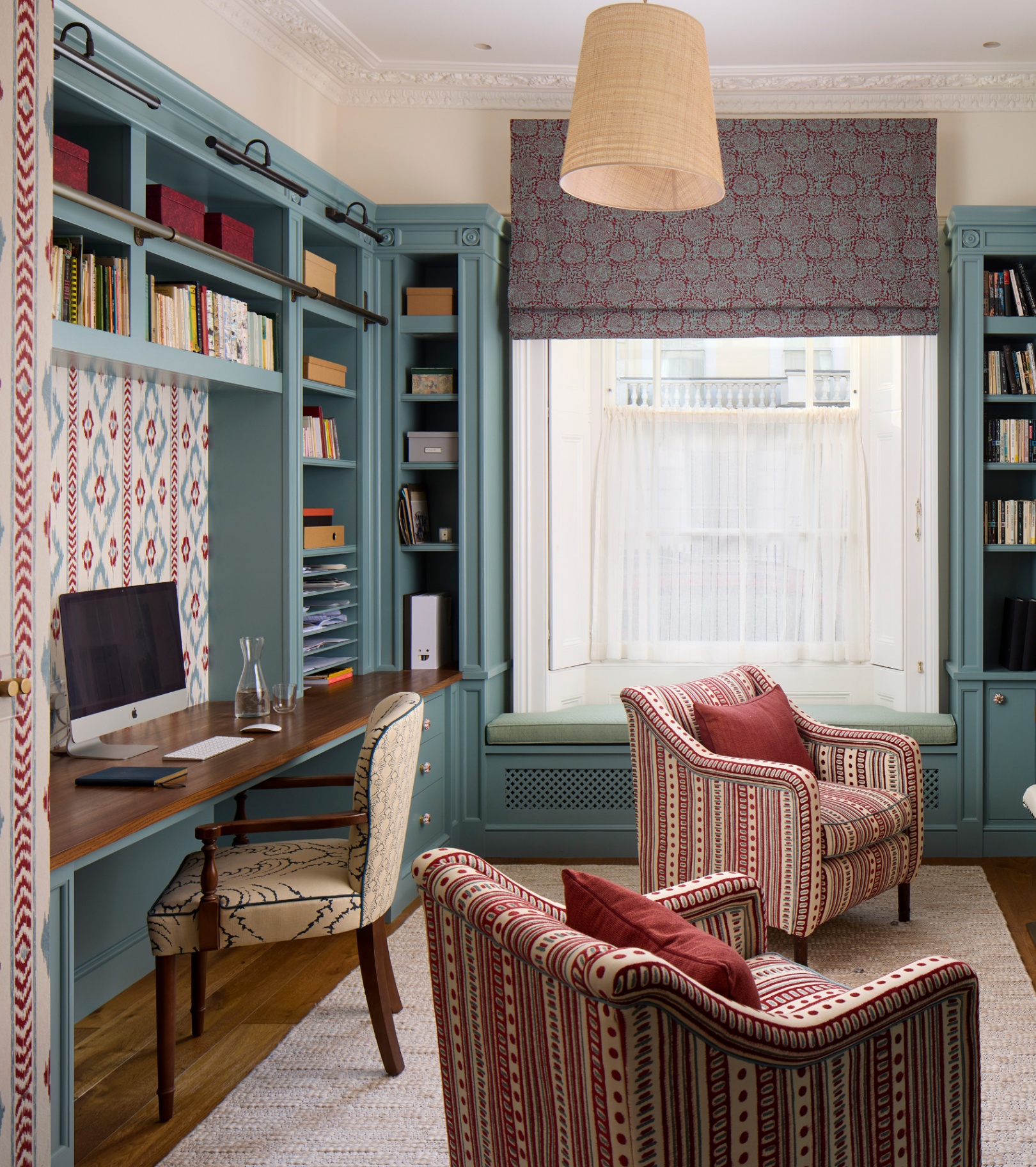 an office with blue painted built in shelving on each wall, a built in desk with a mac on top and a cream upholstered chair