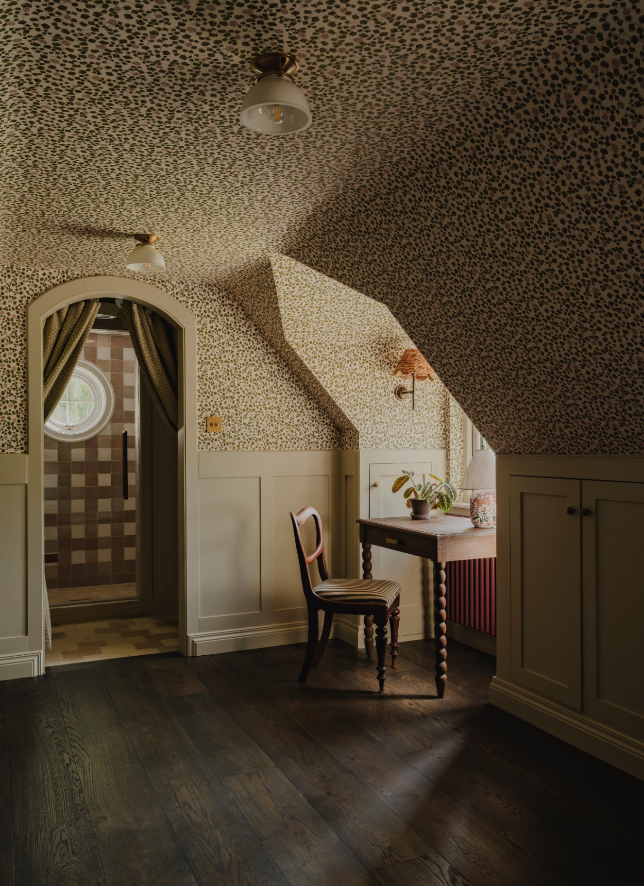 a room with sloped ceilings in a soft beige floral wallpaper and half panelled walls, a small desk and chair under a window and an open door looking through to a an ensuite with brown gingham wallpaper