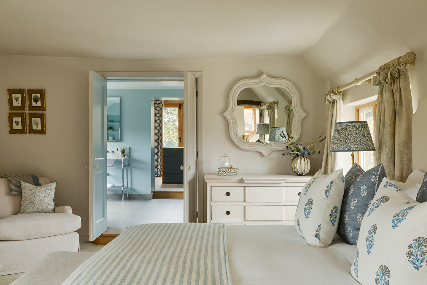 A neutral bedroom with a bed in the foreground and an armchair and chest of drawers with a mirror above on the far wall. An open door looks through to a blue bathroom