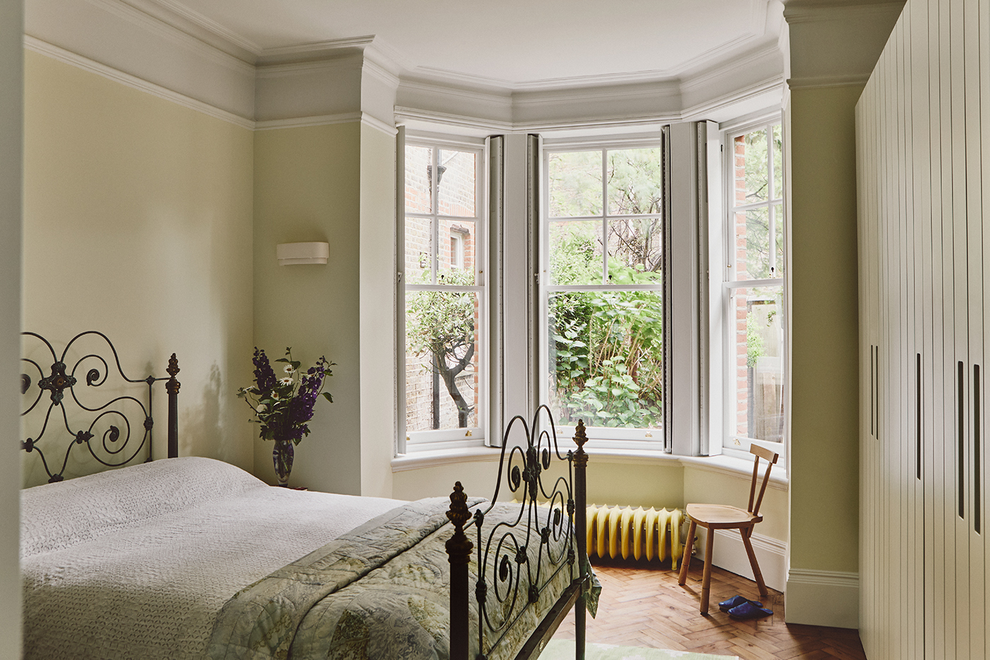 A pale yellow bedroom with high ceilings, a large bay window and wood floor. On the left is a decorative wrought iron double bed dressed with white bedding and a green quilt. On the right are tall cabinets with tongue-and-groove fronts