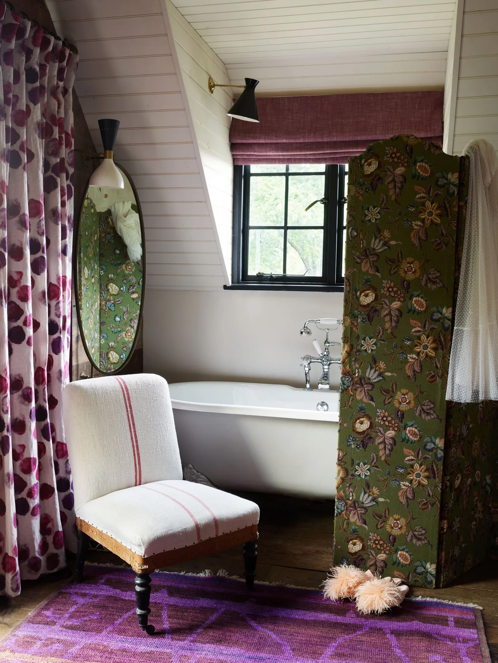 a bathroom with a green floral screen, a pink and purple mat, a cream armchair and a roll top bath