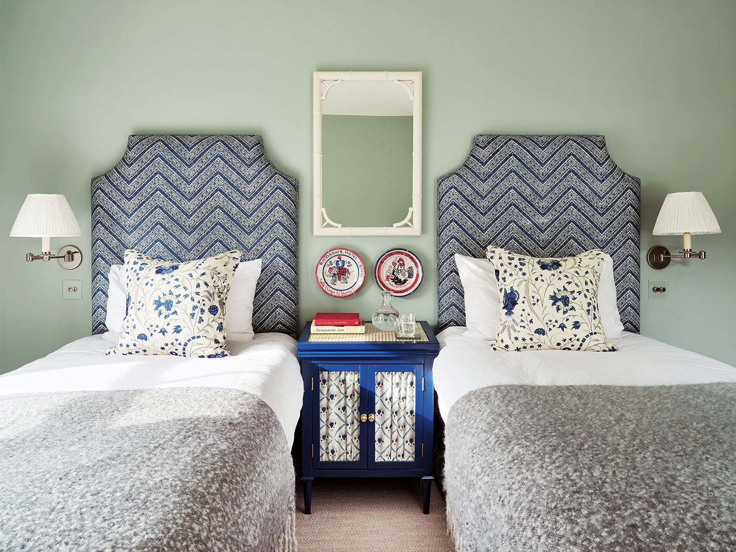 A blue bedroom with twin beds with upholstered patterned blue headboards and dressed with blue floral cushions and grey wool throws. In between sits a blue cabinet with fabric-fronted panels, topped with books and a glass carafe and tumbler