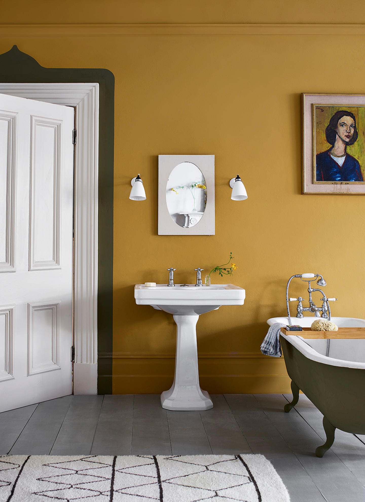 Mustard yellow bathroom with a white pedestal sink, small oval mirror flanked by white sconces, and an olive-green clawfoot bathtub. A portrait hangs on the wall beside an open white door with a decorative dark border outline