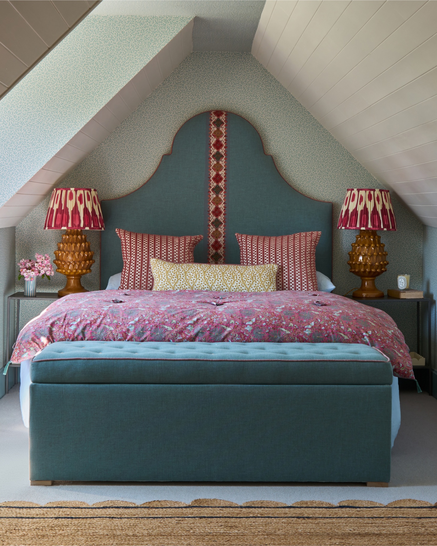 an attic bedroom with white and blue patterned wallpaper, a bed with a large blue and red headboard, two matching lamps either side of the bed and an upholstered storage bench at the end of the bed