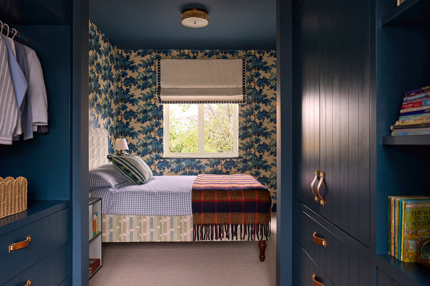 A view through a narrow dressing room area into a bedroom with a blue botanical wallpaper lining the walls and a painted dark blue ceiling. A bed sits in the middle dressed with a tasseled throw and decorative cushion