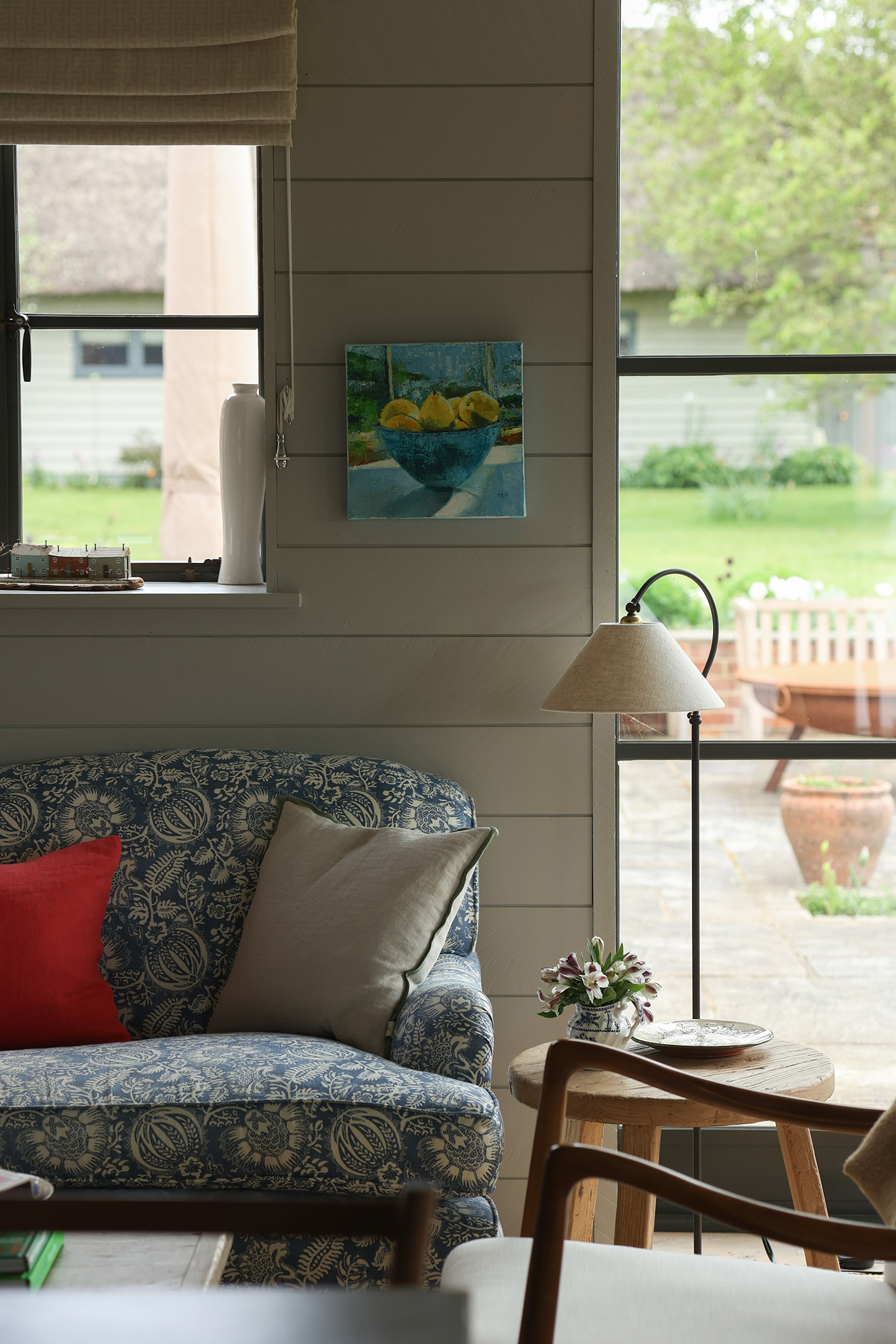 A close up of a blue floral patterned sofa, with a metal table lamp on a wooden side table next to it. On the tongue and groove wall above hangs a painting of lemons in a bowl